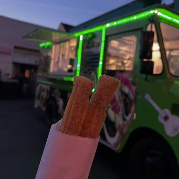 Two churros in a pink wrapper held in front of the Silantro Lime Tacos food truck at dusk.