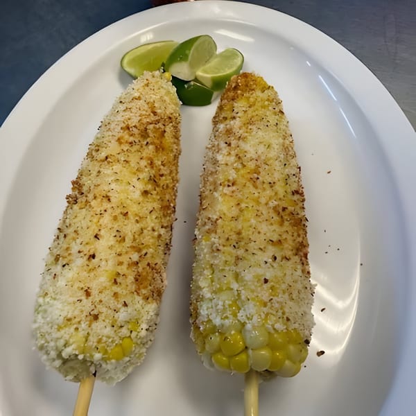 Two Mexican elotes, grilled corn on the cob with cheese and spices, served with lime wedges.