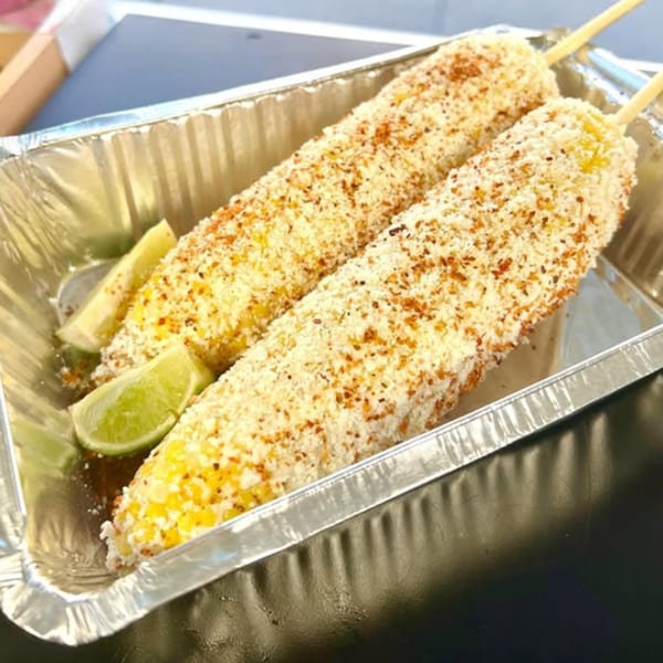 Two elotes, Mexican street corn, covered in cheese and chili powder, with lime wedges in a foil tray.