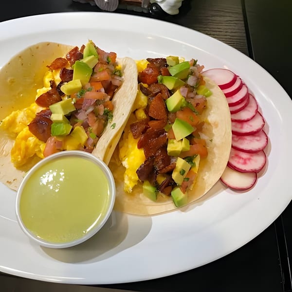Two breakfast tacos with scrambled eggs, bacon, avocado, pico de gallo, and a side of radishes.