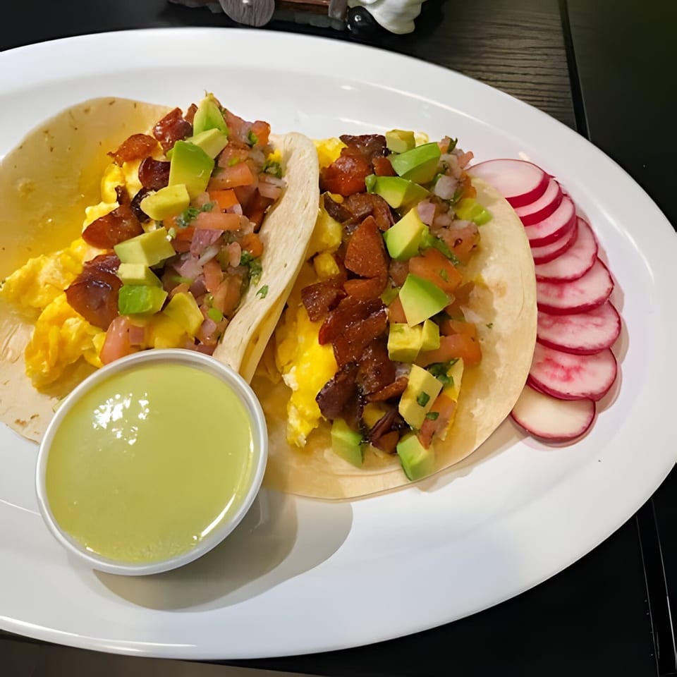 Two breakfast tacos with scrambled eggs, bacon, avocado, pico de gallo, and a side of radishes.